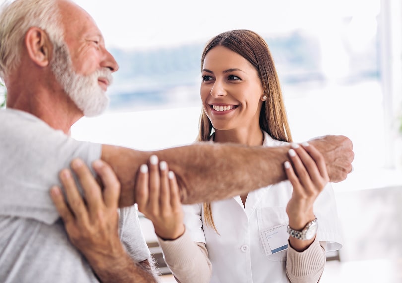 A healthcare professional assists an elderly man with an arm exercise in a bright setting.