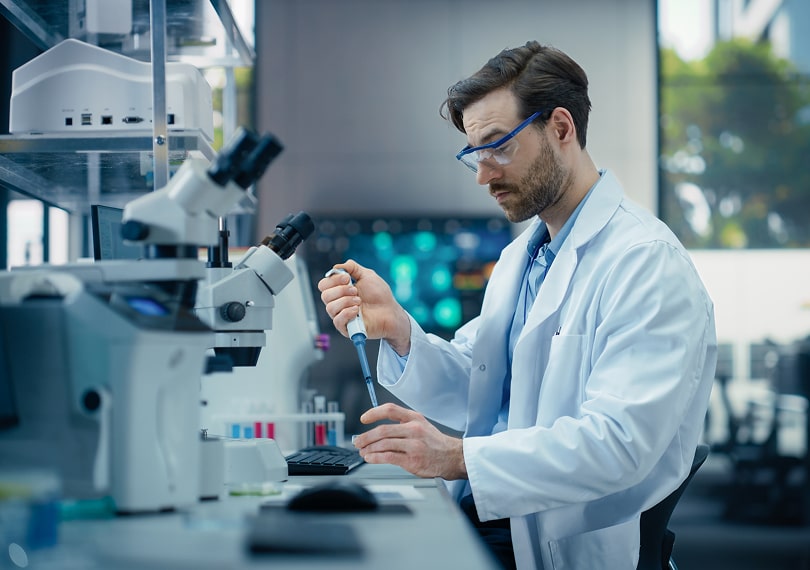 Scientist in a lab coat uses a pipette beside a microscope in a laboratory setting.