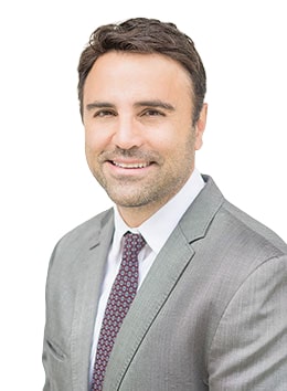 Man wearing a light gray suit, white shirt, and patterned tie, smiling at the camera against a plain white background.