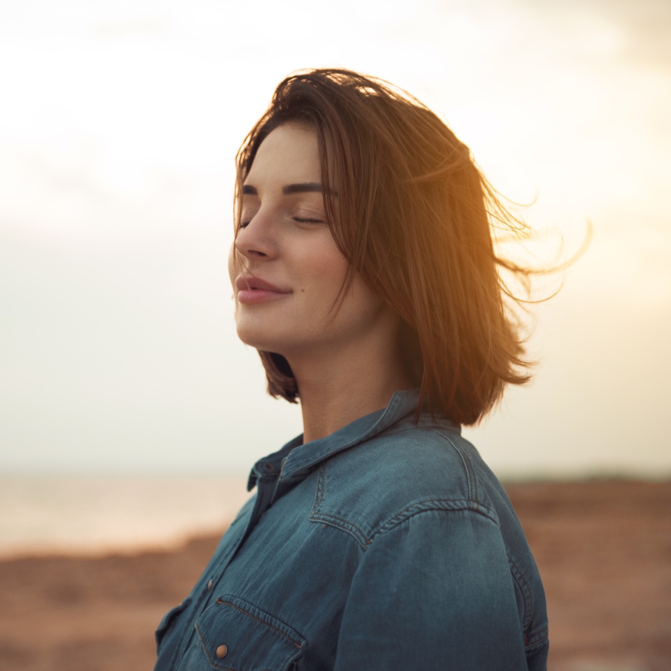 Great pleasure. Charming calm young woman is standing near sea with closed eyes and expressing delight. She is posing against wonderful sunset while enjoying last rays of the sun