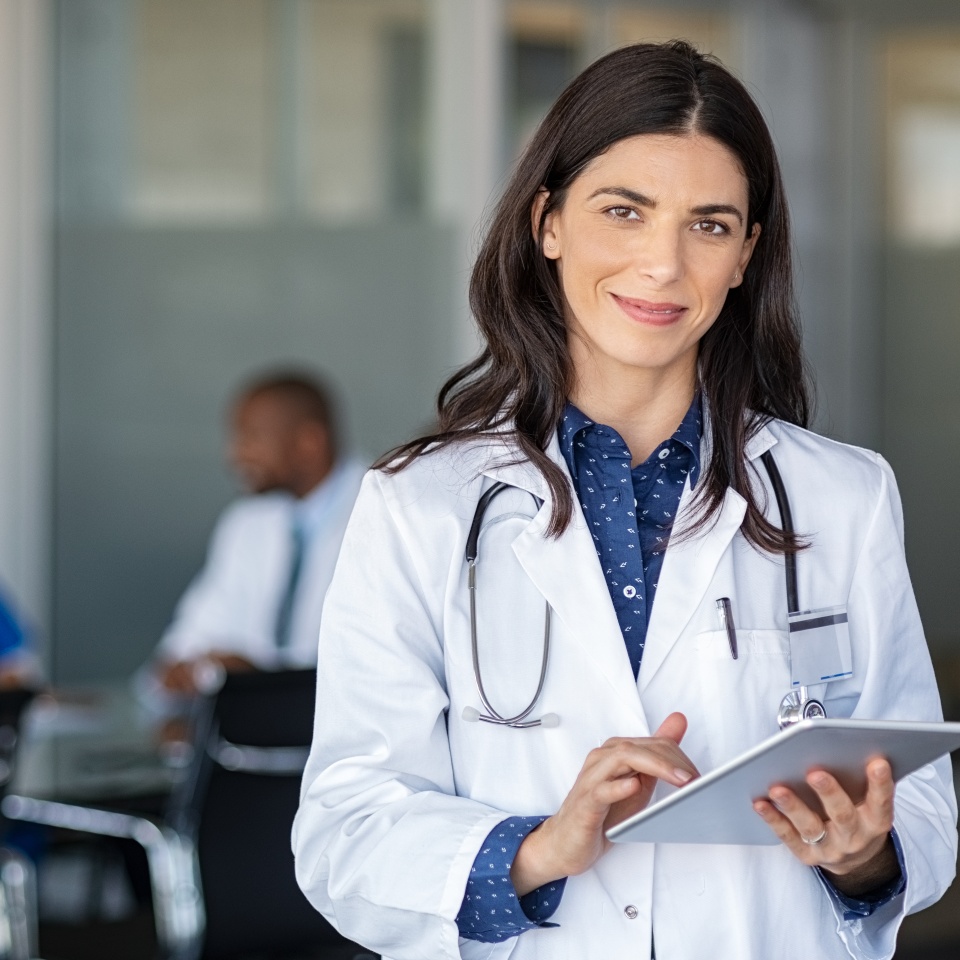Portrait of beautiful mature woman doctor holding digital tablet and looking at camera. Confident female doctor using digital tablet with colleague talking in background at hospital. Latin nurse.
