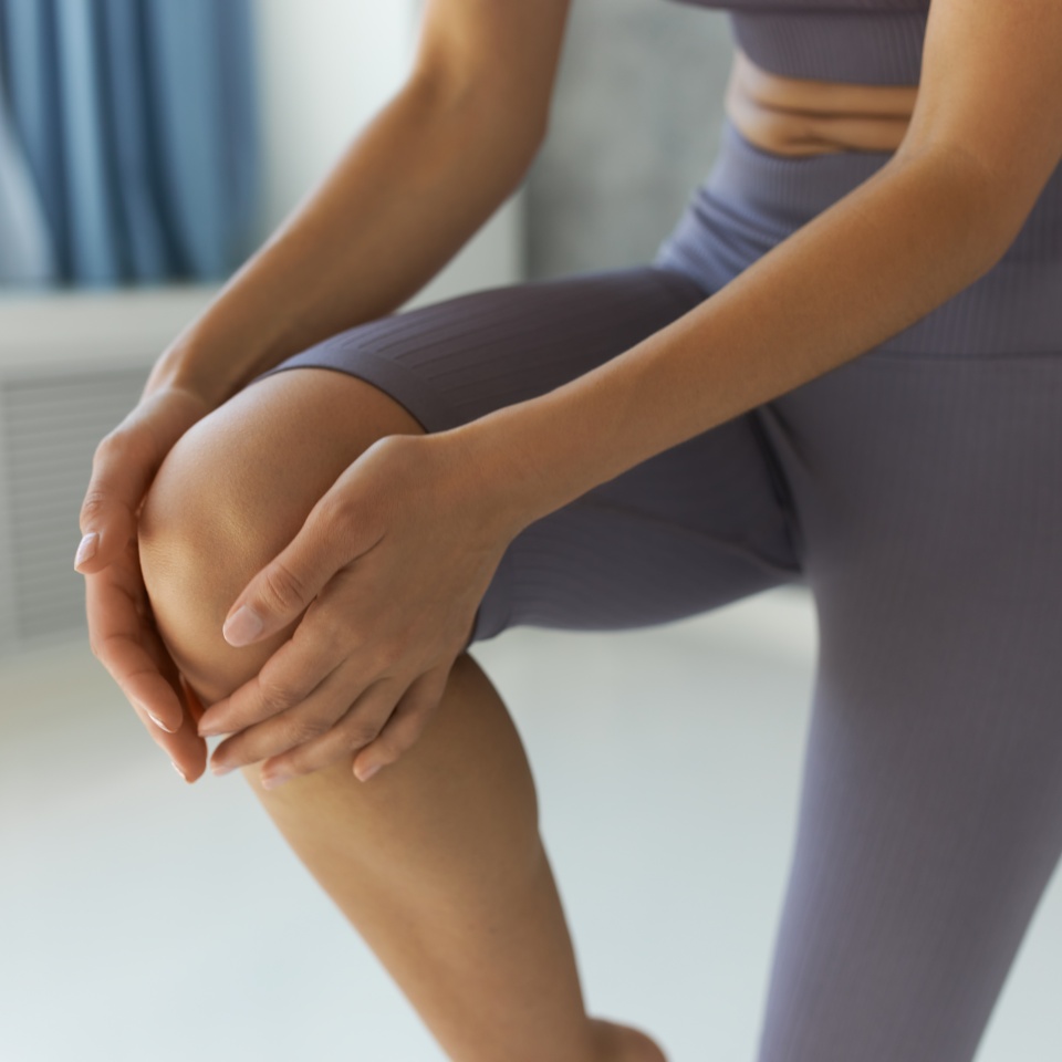 Crop shot of random Caucasian woman doing yoga in lilac crop top and leggings, touching gently right knee, balancing on left foot, having cute abdominal folds, isolated over fitness studio background