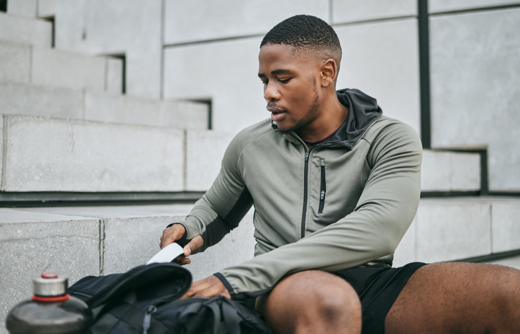 A man in athletic clothing sits on outdoor concrete steps, looking into his gym bag with a water bottle nearby.