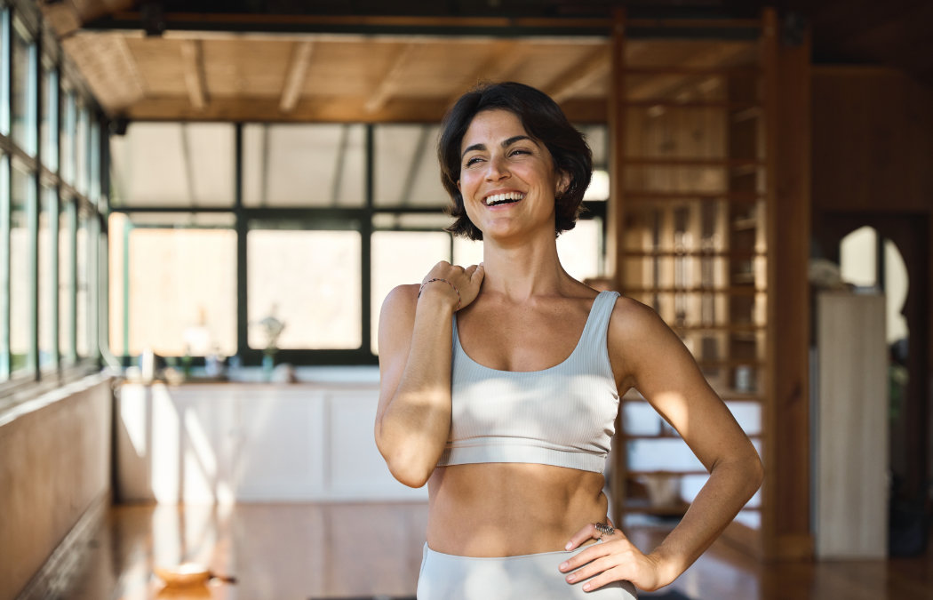 Woman in athletic wear smiles and stands in a sunlit room with wooden floors and large windows.