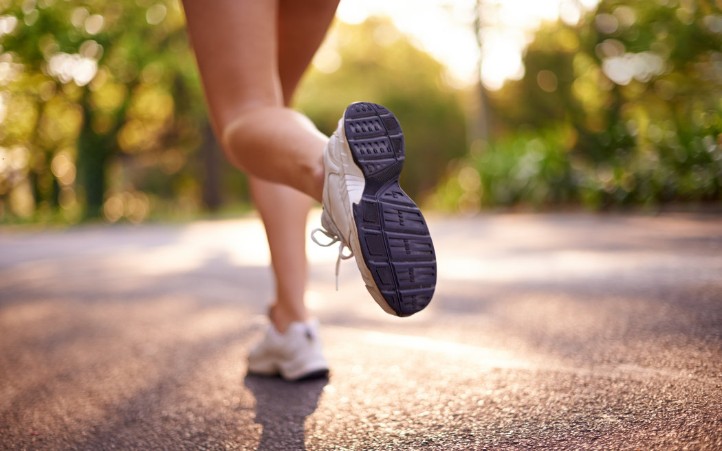 Close-up of a person running outdoors on a paved path, focusing on the sole of one athletic shoe with blurred trees in the background.