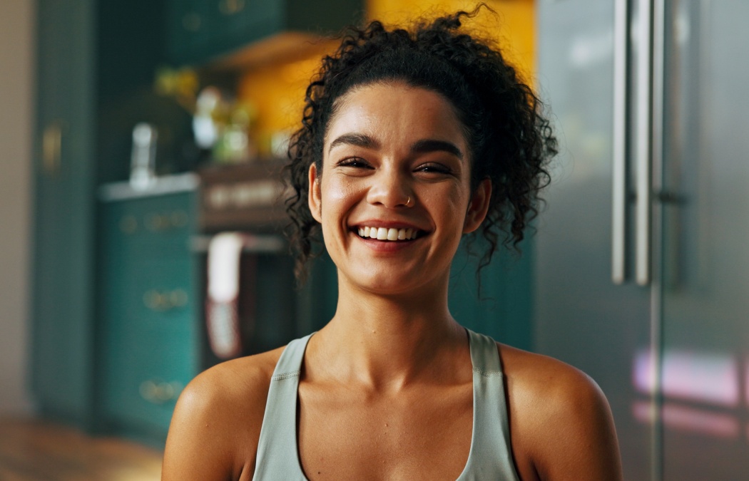 A woman with curly hair in a tank top smiles at the camera indoors, with a kitchen visible in the background.